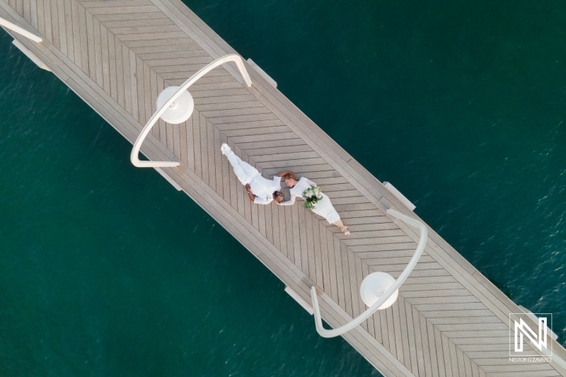 Aerial view of a romantic wedding couple lying on a wooden dock over turquoise water during sunset, surrounded by soft lighting and a serene atmosphere