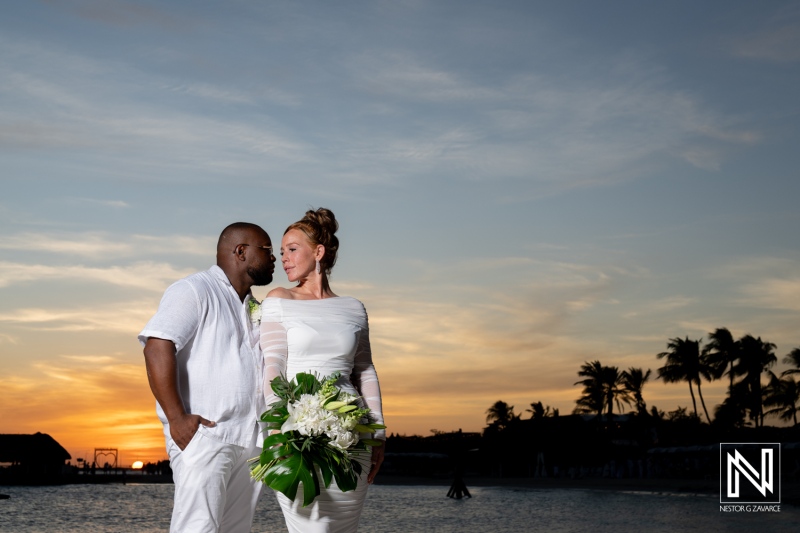 A couple joyfully embraces during their beach wedding ceremony at sunset, surrounded by palm trees and ocean waves beneath a colorful sky