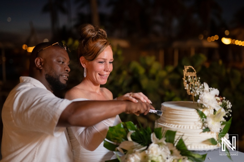 Couple joyfully cutting their wedding cake during a romantic outdoor celebration at sunset in a beautiful tropical setting