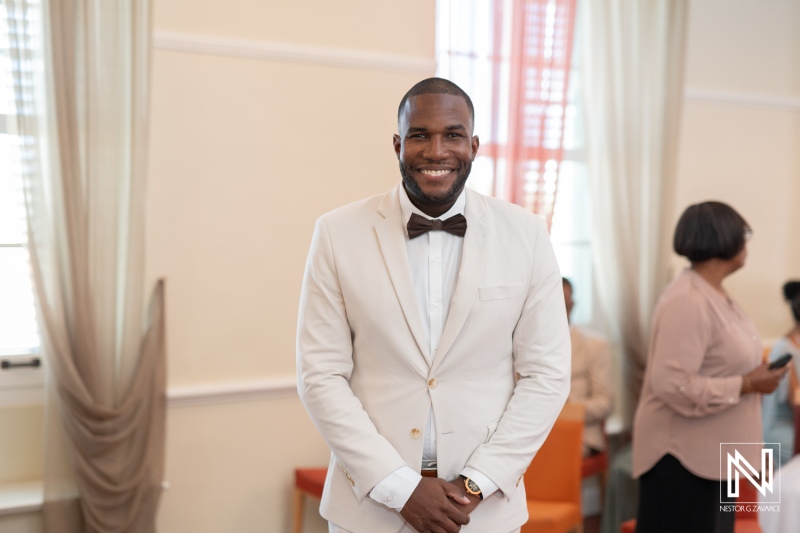 A smiling man in a light suit with a bow tie stands confidently in a well-lit room during a formal event, surrounded by guests engaging in conversation