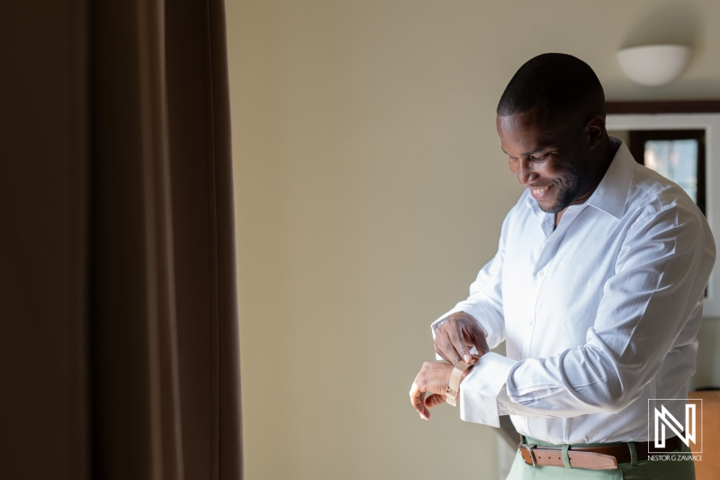 A groom prepares for his wedding day in a bright hotel room while adjusting his cufflinks with a joyful smile on his face during a beautiful morning