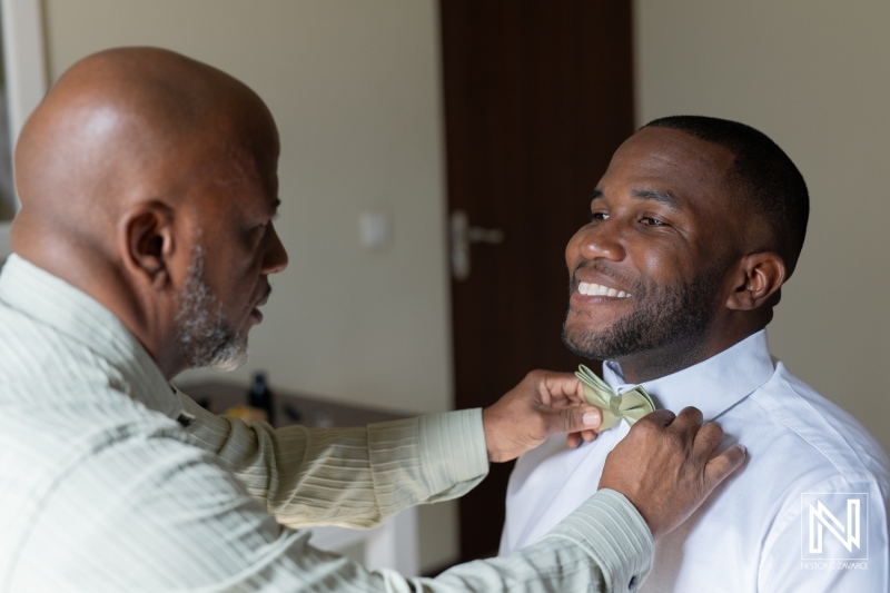 A father adjusts his son\'s bow tie while getting ready for a wedding in a hotel room during the morning hours