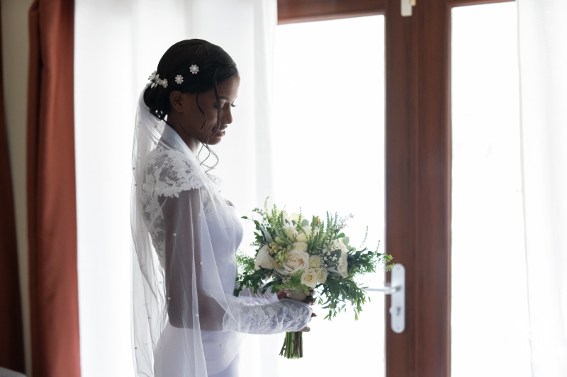 A bride stands by a window holding her bouquet while preparing for her wedding ceremony in a softly lit room on a sunny day