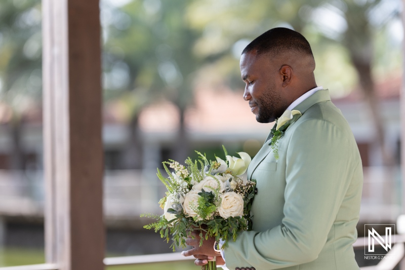 A groom stands quietly with a bouquet of white flowers, taking a moment before the wedding ceremony at a scenic outdoor venue on a sunny day