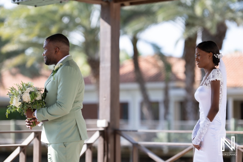 A joyful outdoor wedding ceremony at a beachfront venue with a groom in green suit and bride in elegant white dress standing together in anticipation of their vows