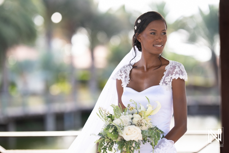 Elegant bride in white gown with bouquet poses outdoors in a serene setting surrounded by palm trees during golden hour