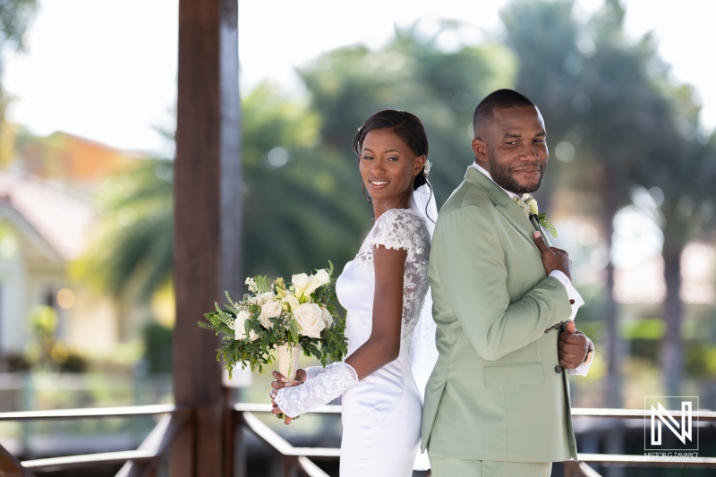 A joyful couple stands back to back, smiling as they celebrate their wedding day in a scenic outdoor venue surrounded by lush greenery and natural light