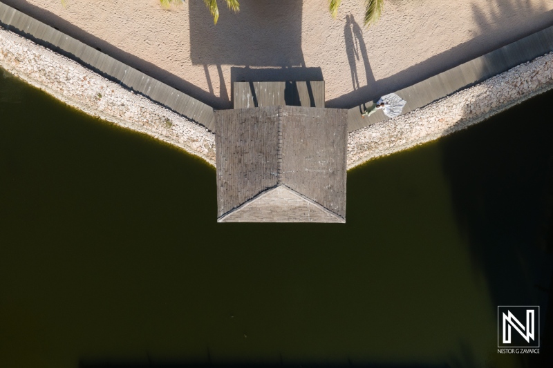 Aerial view of a small wooden dock extending into a dark green lake surrounded by sandy beach in a serene coastal location, during bright sunlight on a clear day