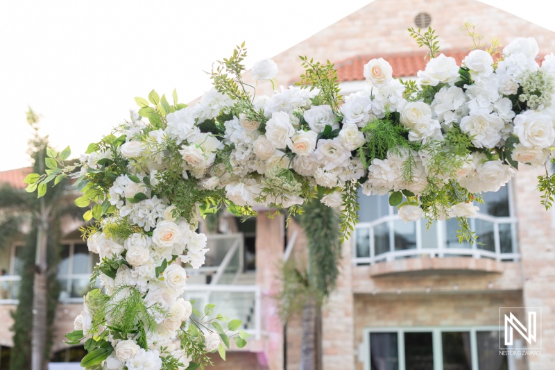 A beautiful floral arch adorned with white roses and greenery sits outside a charming venue, ready for a wedding ceremony during golden hour