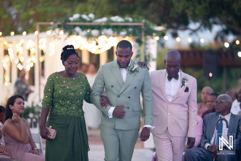 A groom accompanied by his parents walks down the aisle during a beautiful outdoor wedding ceremony at sunset with elegant decorations and joyful guests