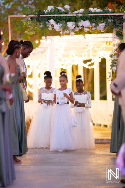 Three flower girls in elegant white dresses walk down the aisle during a wedding ceremony in a beautifully decorated outdoor venue during the evening