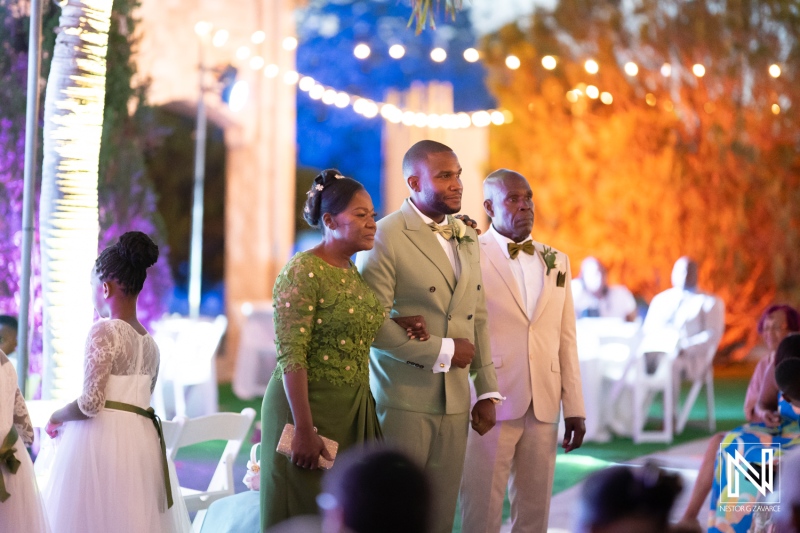 A wedding ceremony featuring a groom in a light suit, flanked by his mother and father, surrounded by guests enjoying the joyful celebration during the evening