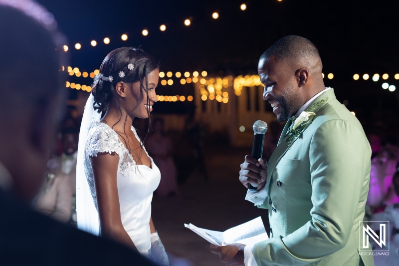 A joyful wedding ceremony under twinkling lights, as the bride smiles at the groom during the heartfelt vows exchanged at sunset in a picturesque outdoor setting