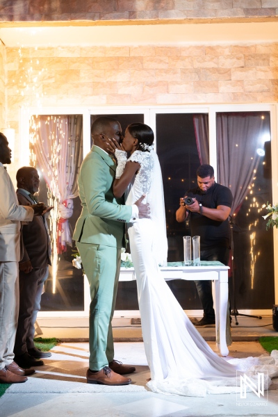 A romantic wedding kiss between a couple in elegant attire at an outdoor venue illuminated with fairy lights during a nighttime celebration