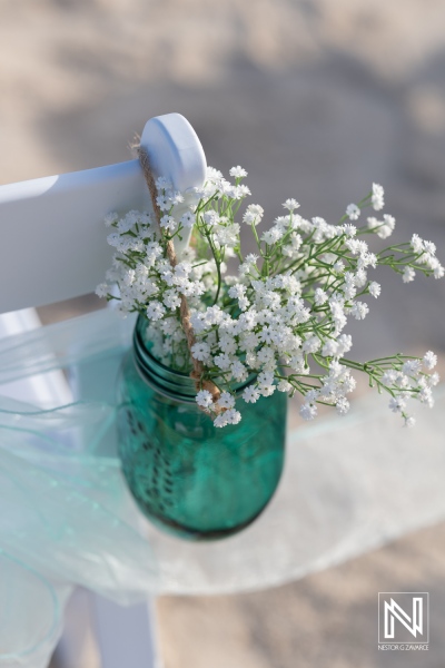 Delicate White Flowers Arranged in a Teal Mason Jar Hanging on a Charming Wooden Chair at an Outdoor Event During Golden Hour Near a Beautiful Venue