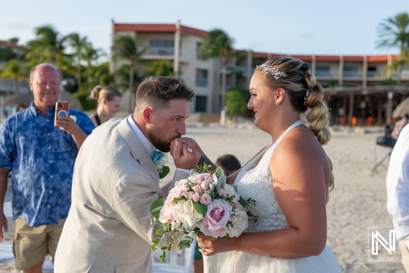 A Groom Kisses the Bride\'s Hand During a Beachfront Wedding Ceremony at Sunset in an Idyllic Tropical Location Surrounded by Family and Friends