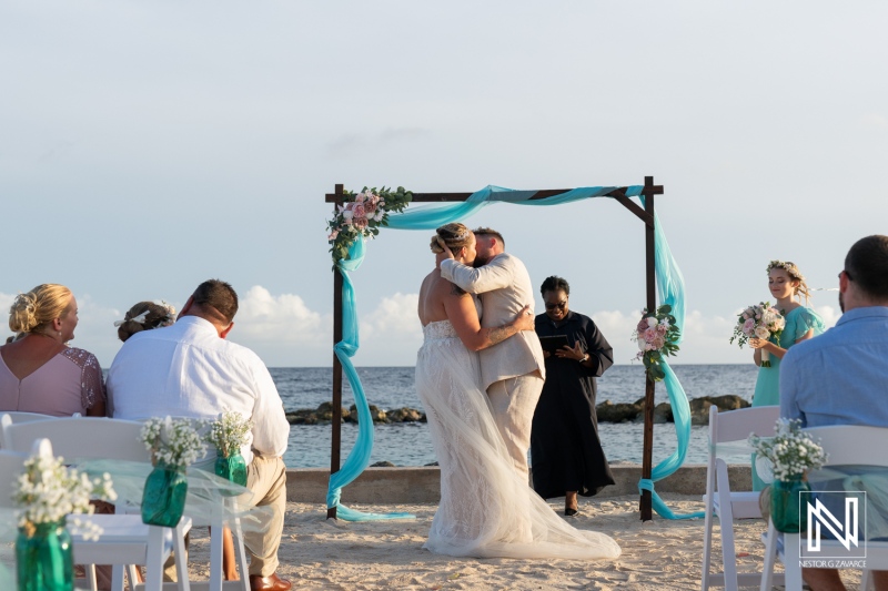 A Beachfront Wedding Ceremony Captures a Couple Sharing Their First Kiss as Newlyweds Under a Floral Altar at Sunset, Surrounded by Family and Friends