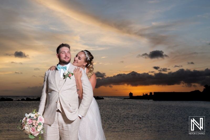 A Happy Newlywed Couple Stands by the Ocean at Sunset, Embracing Warmly as the Sky Glows With Soft Colors in This Romantic Wedding Moment