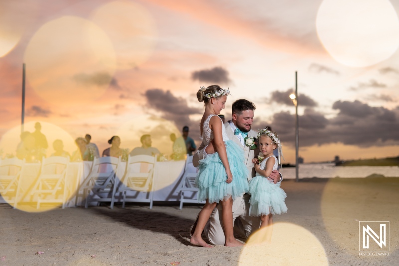 A Joyful Father Embraces His Daughters Dressed in Matching Light Blue Dresses During a Vibrant Sunset Celebration by the Beach, Capturing a Moment of Love and Happiness