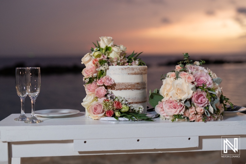 A Beautifully Arranged Wedding Cake and Floral Bouquet on a White Table at Sunset by the Beach, Capturing a Romantic Celebration in a Serene Coastal Setting