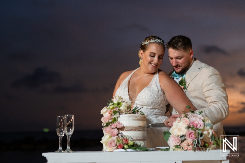 A Joyful Couple Cuts Their Elegant Wedding Cake on a Beach at Sunset, Surrounded by Floral Arrangements and Champagne Glasses