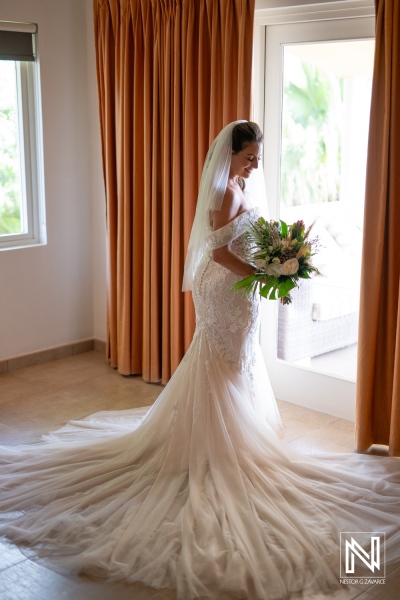 A Bride in a Stunning Gown Stands Near a Window, Holding a Bouquet of Flowers, Preparing for Her Wedding Day in an Elegant Location Filled With Natural Light