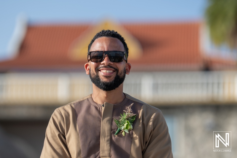 A Smiling Man in Sunglasses Showcases a Stylish Outfit With a Floral Accent During a Daytime Outdoor Celebration Near a Historic Venue