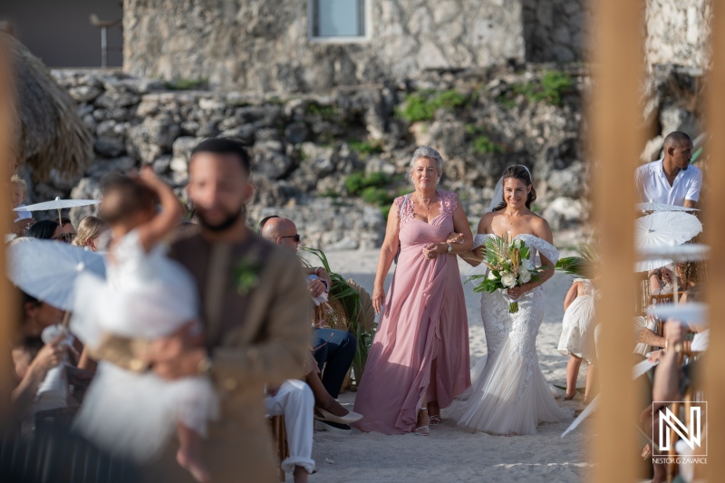 A Beautiful Beach Wedding Ceremony Taking Place at Sunset, With the Bride and Her Mother Walking Down the Aisle in a Tropical Coastal Setting Surrounded by Guests