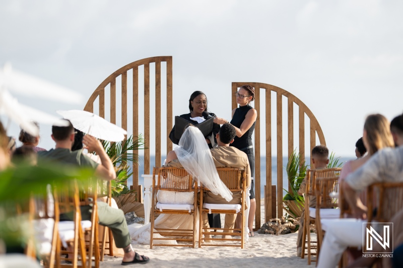 A Beachfront Wedding Ceremony Takes Place Under a Wooden Arch on a Sunny Day, With Guests Seated in Bamboo Chairs, Enjoying a Tropical Breeze and Scenic Ocean Backdrop