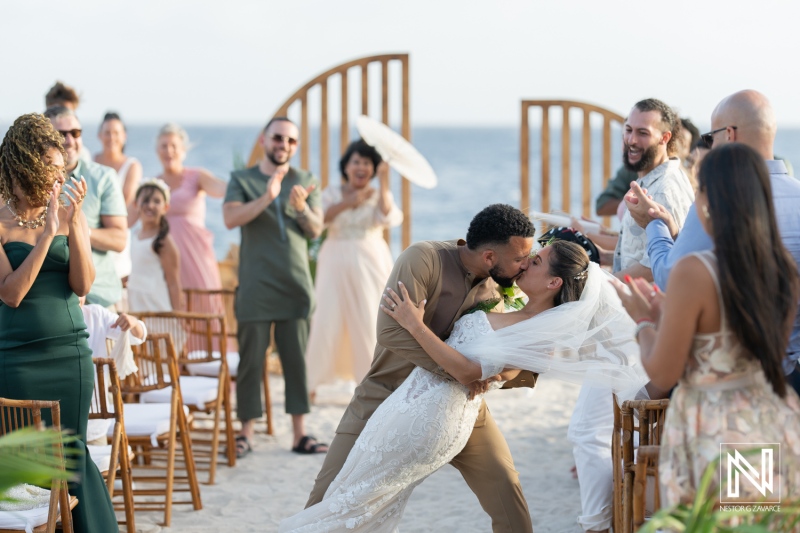 Couple Shares Their First Kiss as a Married Pair on a Picturesque Beach During a Sunset Wedding Ceremony Surrounded by Joyful Friends and Family