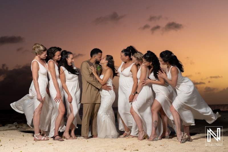 A Joyful Couple Shares a Romantic Kiss on the Beach, Surrounded by Bridesmaids Enjoying the Sunset in a Beautiful Tropical Setting