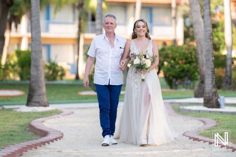 Bride walks down sandy path with her father, adorned in a beautiful white gown, as they celebrate a joyful moment during an outdoor wedding in a tropical resort