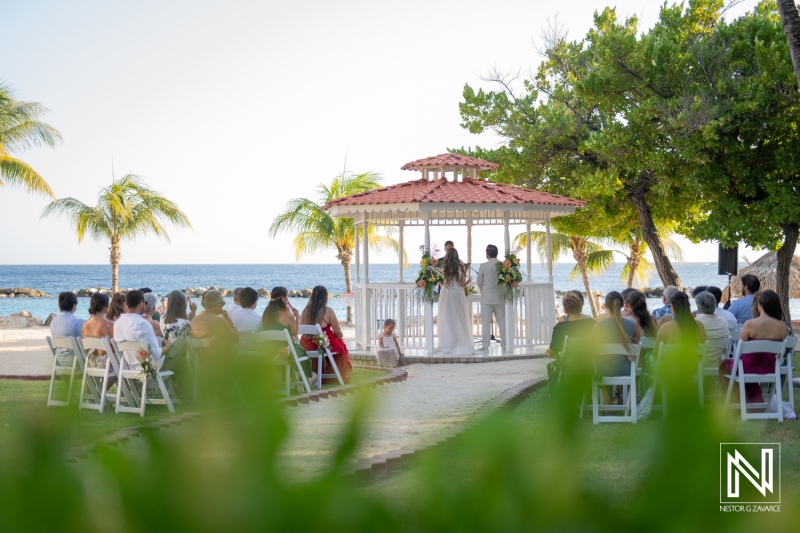 A beautiful beach wedding ceremony takes place under a gazebo surrounded by palm trees and guests, capturing a romantic moment by the sea in daylight