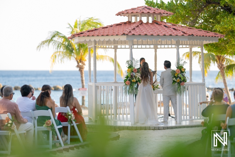 A beautiful beach wedding ceremony at a gazebo under a blue sky with palm trees, surrounded by friends and family during sunset