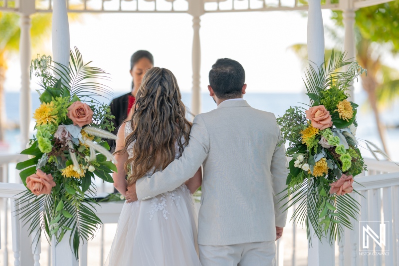 A couple exchanges vows during a beach wedding ceremony with floral decorations under a gazebo near the ocean at sunset