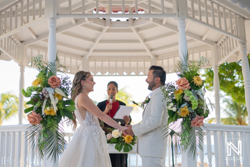 A beautiful beach wedding ceremony takes place under a floral gazebo with a joyful couple exchanging vows at sunset by the ocean