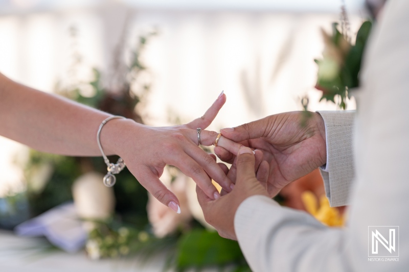 A couple exchanges wedding rings during a beautiful outdoor ceremony surrounded by flowers and nature in a sunny setting