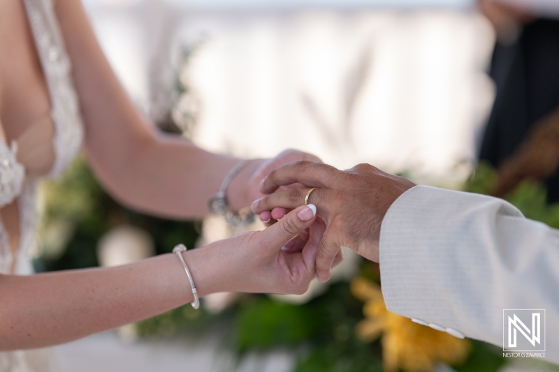 Wedding couple exchanging rings during a beach ceremony at sunset, surrounded by flowers and greenery, celebrating their love and commitment in a beautiful outdoor setting