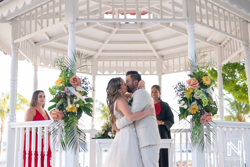 Couple shares a romantic kiss during their outdoor wedding ceremony at a beautifully decorated gazebo by the beach at sunset