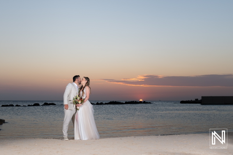 A couple shares a romantic kiss at sunset on a serene beach during their wedding ceremony by the ocean, surrounded by soft waves and vibrant clouds