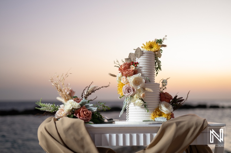 Elegant wedding cake adorned with flowers displayed on a table at sunset by the beach with soft colors of the horizon highlighting the moment