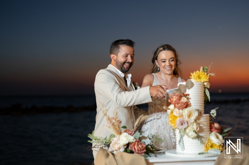 A couple joyfully cutting their wedding cake at sunset by the beach in a romantic coastal setting with colorful floral decorations