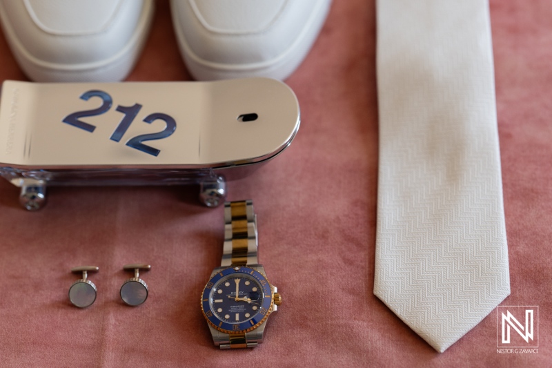 Wedding accessories arranged neatly on a blush tablecloth featuring shoes, a tie, cufflinks, a watch, and a decorative sign in Curacao