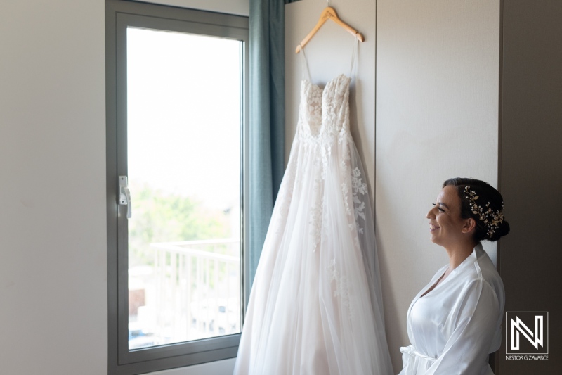 Bride preparing for her wedding day in Curacao with a beautiful dress hanging by the window, capturing a moment of anticipation and joy