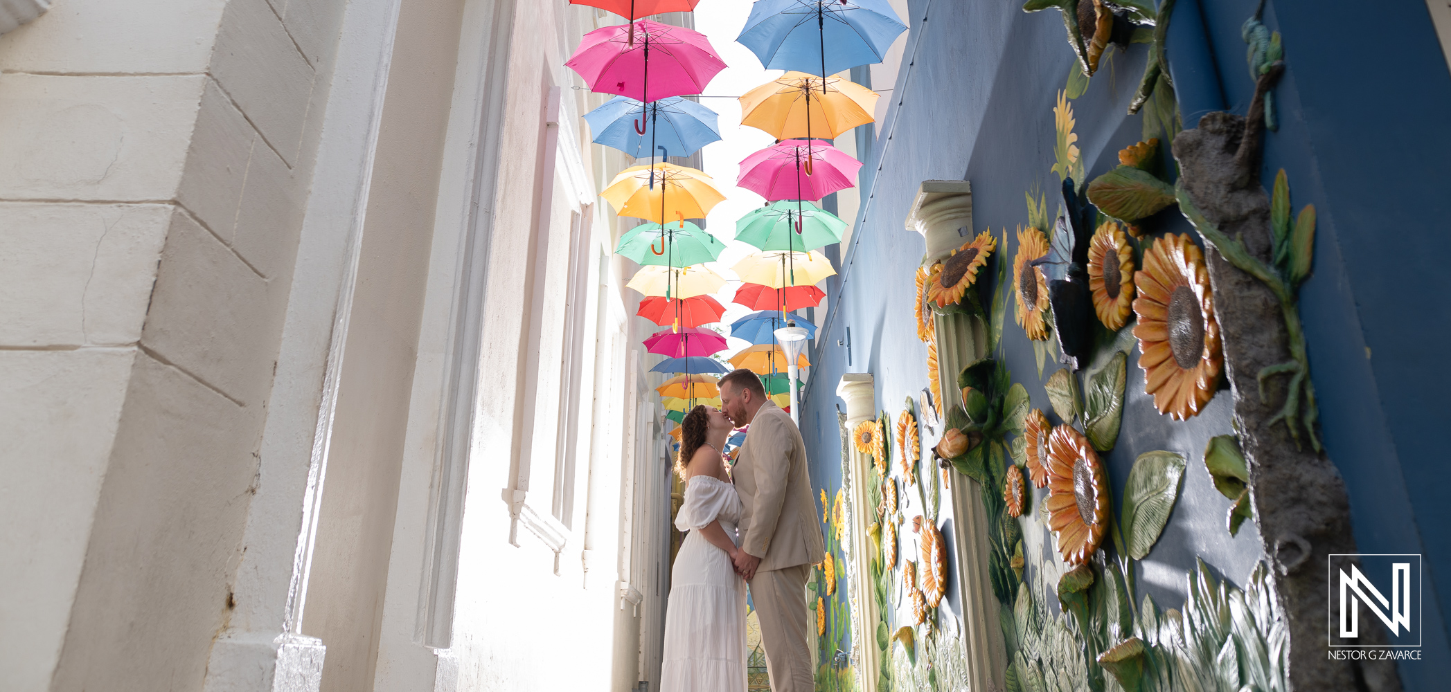 Julia and James pose beneath colorful hanging umbrellas beside a decorated blue wall in Punda during their Curacao trash the dress session.
